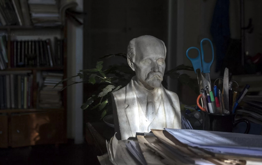 Portreta skulptūra, kura atrodas uz darba galda starp papīriem, kancelejas rīkiem un grāmatām