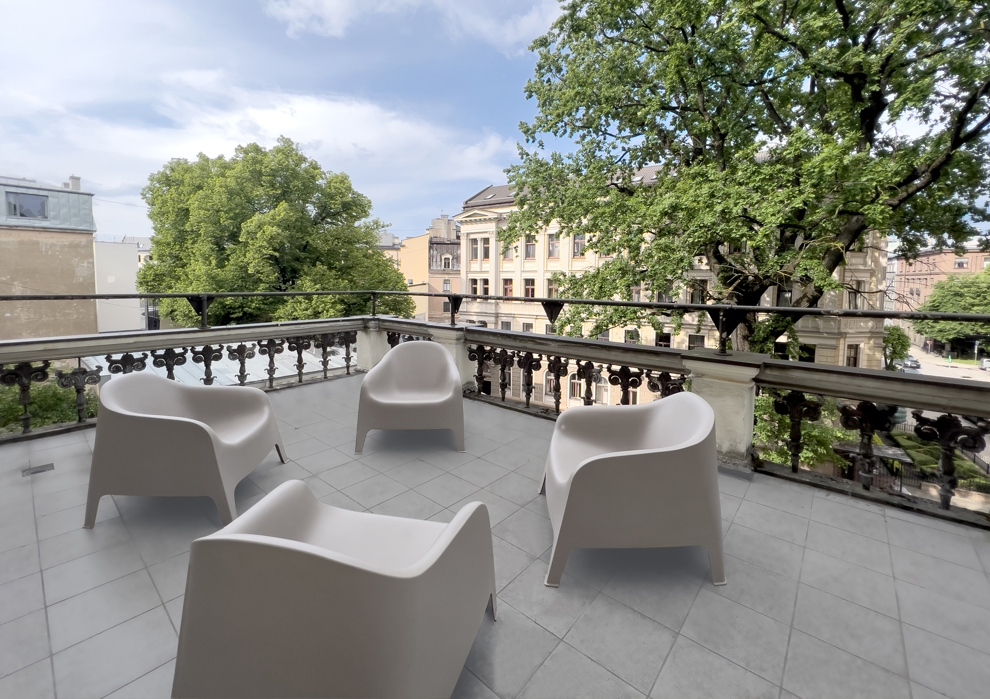 Image of the third-floor balcony. Light grey tiled floor, four sand-colored lounge chairs in the center of the balcony. In the background, a sand-colored residential building across the street. In the foreground, two lush green trees behind the balcony.