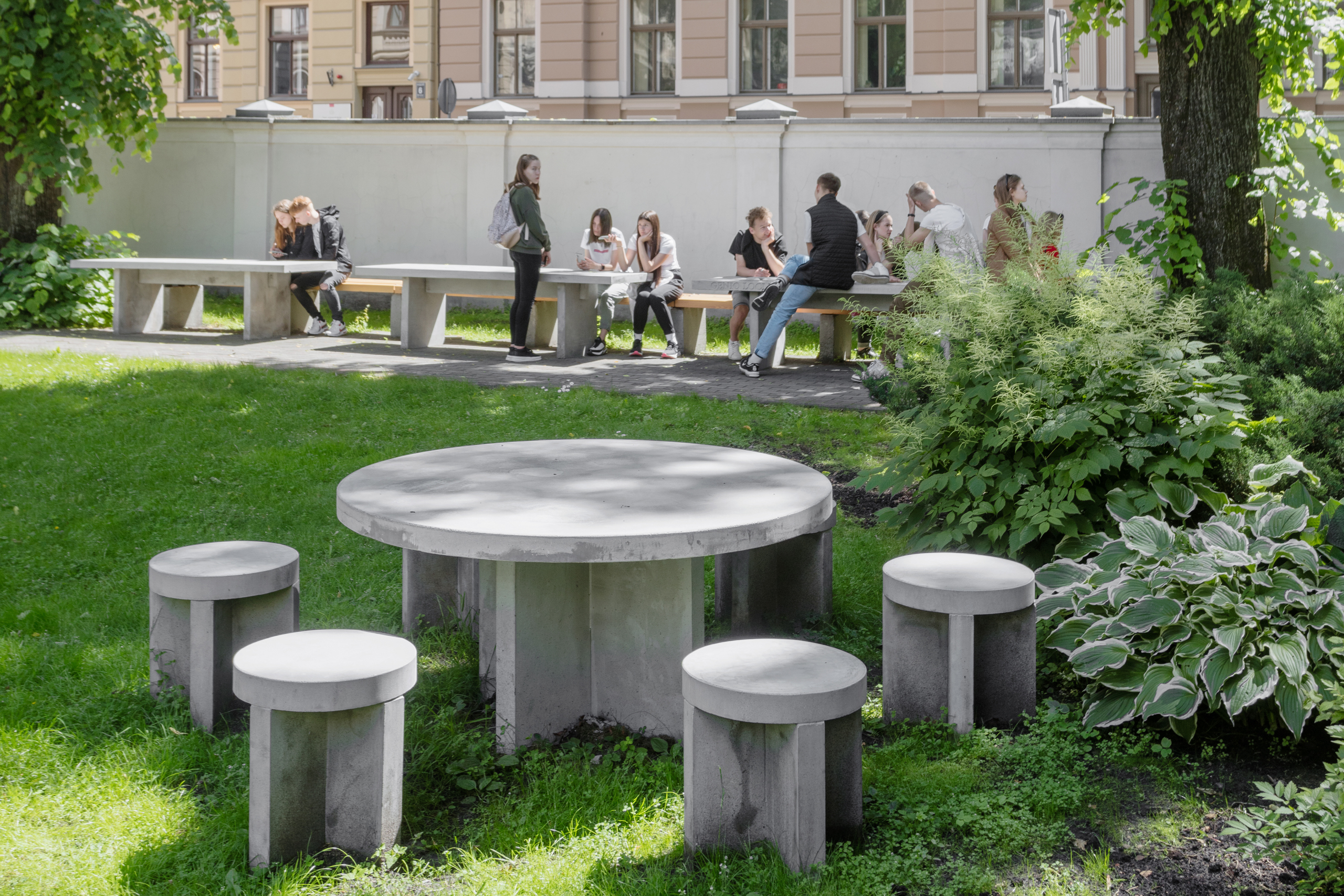 Museum garden. In the foreground is a circular concrete table with four chairs around it. In the background, green grass with trees and people sitting on concrete garden benches and tables.