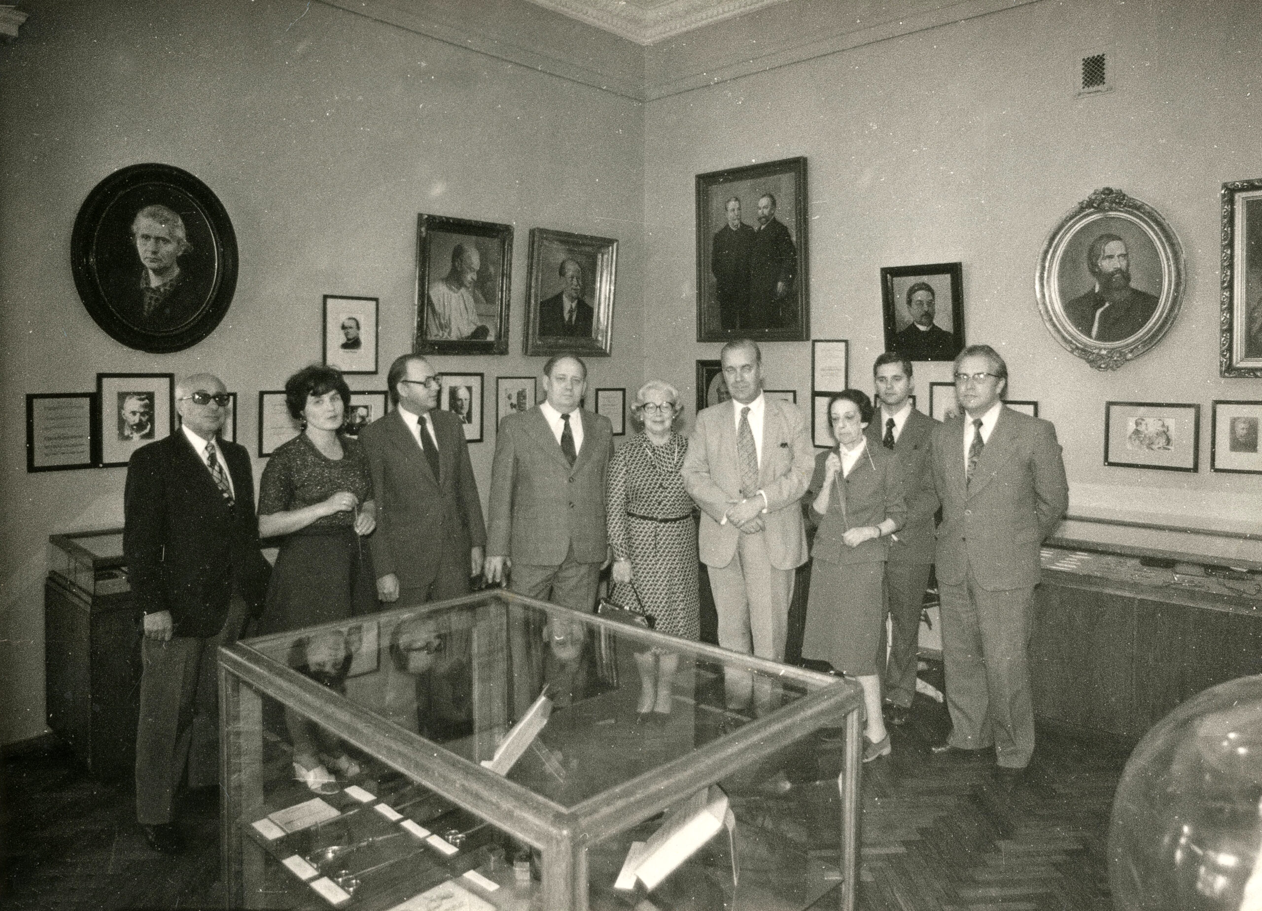 Nine people have lined up in the middle of one of the exhibition halls of the museum. A glass display case can be seen in the foreground. In the background there are other showcases and on the wall you can see many different paintings and photographs, mostly portraits.
