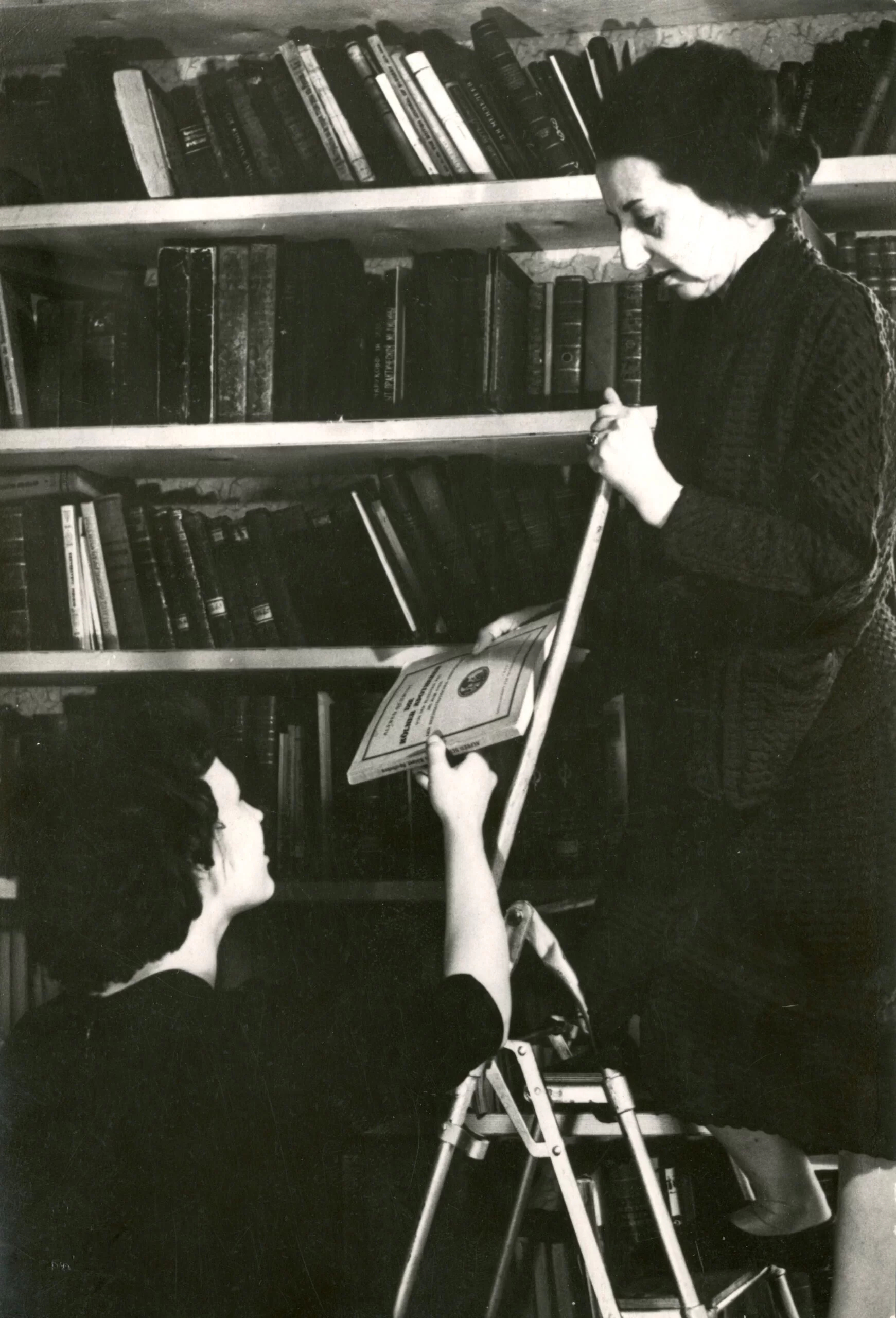 Photo from the museum library. Two women can be seen, one climbing the stairs and handing the book to the other. In the background is a large shelf full of books.