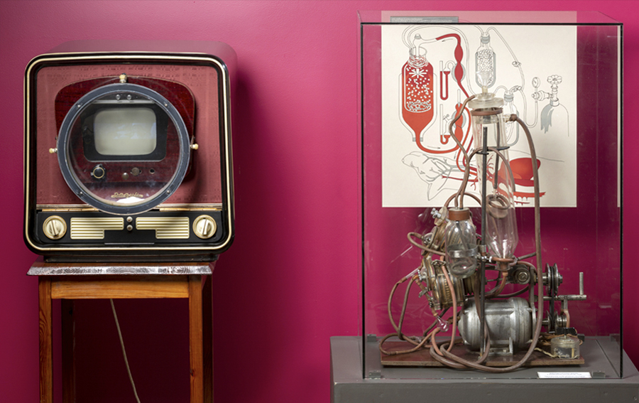 Fotogrāfija no muzeja ekspozīcijas ar vēsturisku televizoru, kam blakus atrodas mākslīgās asinsrites aparāts.